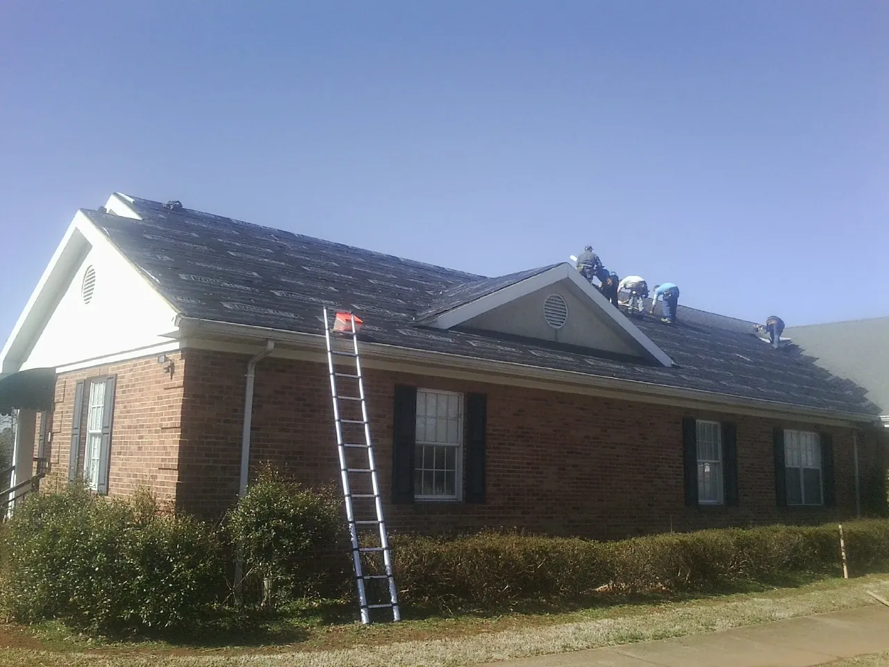 Crew tearing shingles off a brick ranch roof with a ladder set — Emergency Roof Repair work by our crew in Mauldin