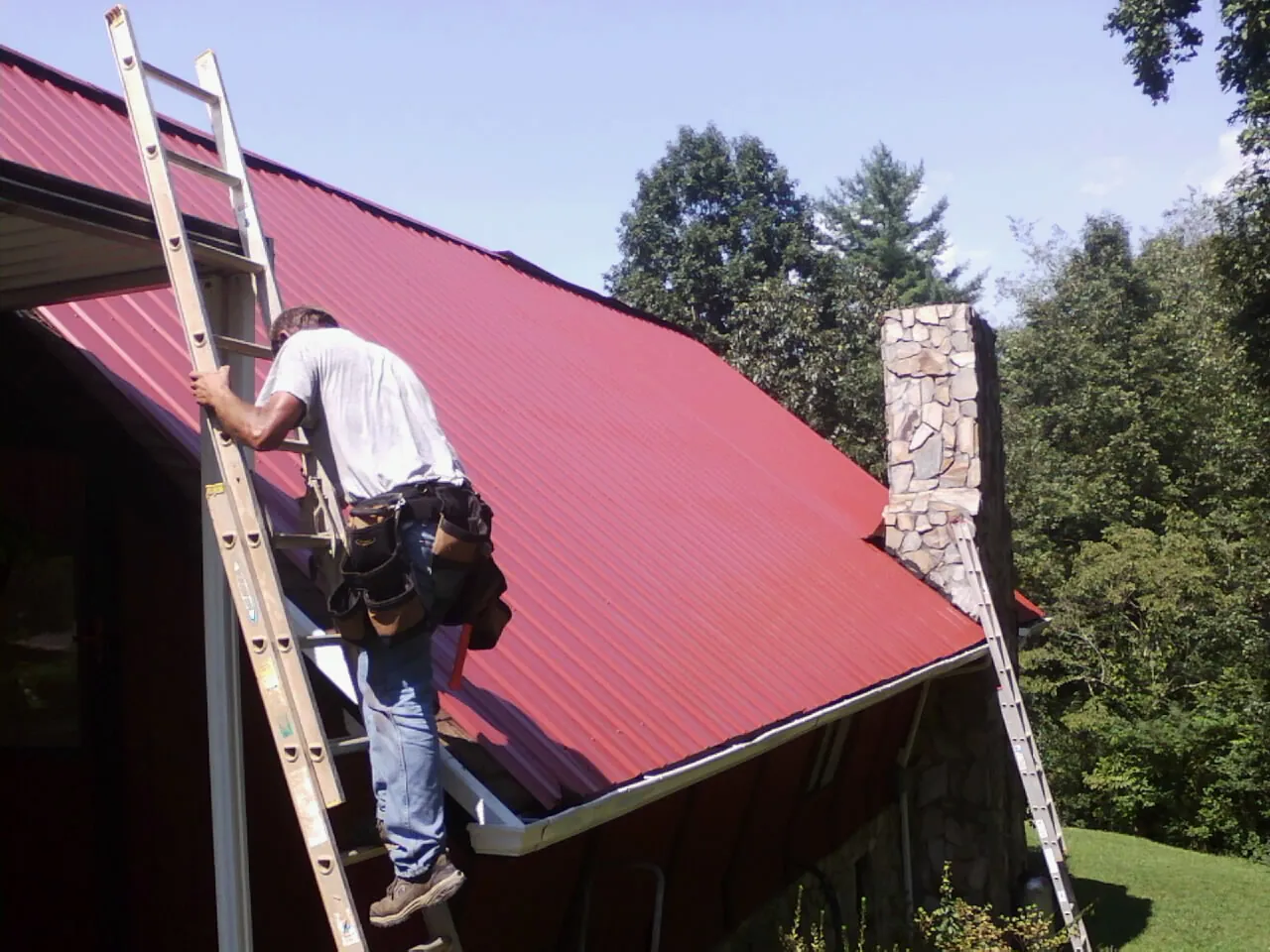 Roofer on a ladder beside a red metal roof and stone chimney — Soffit & Fascia Repair by Roofing Brothers in Mauldin